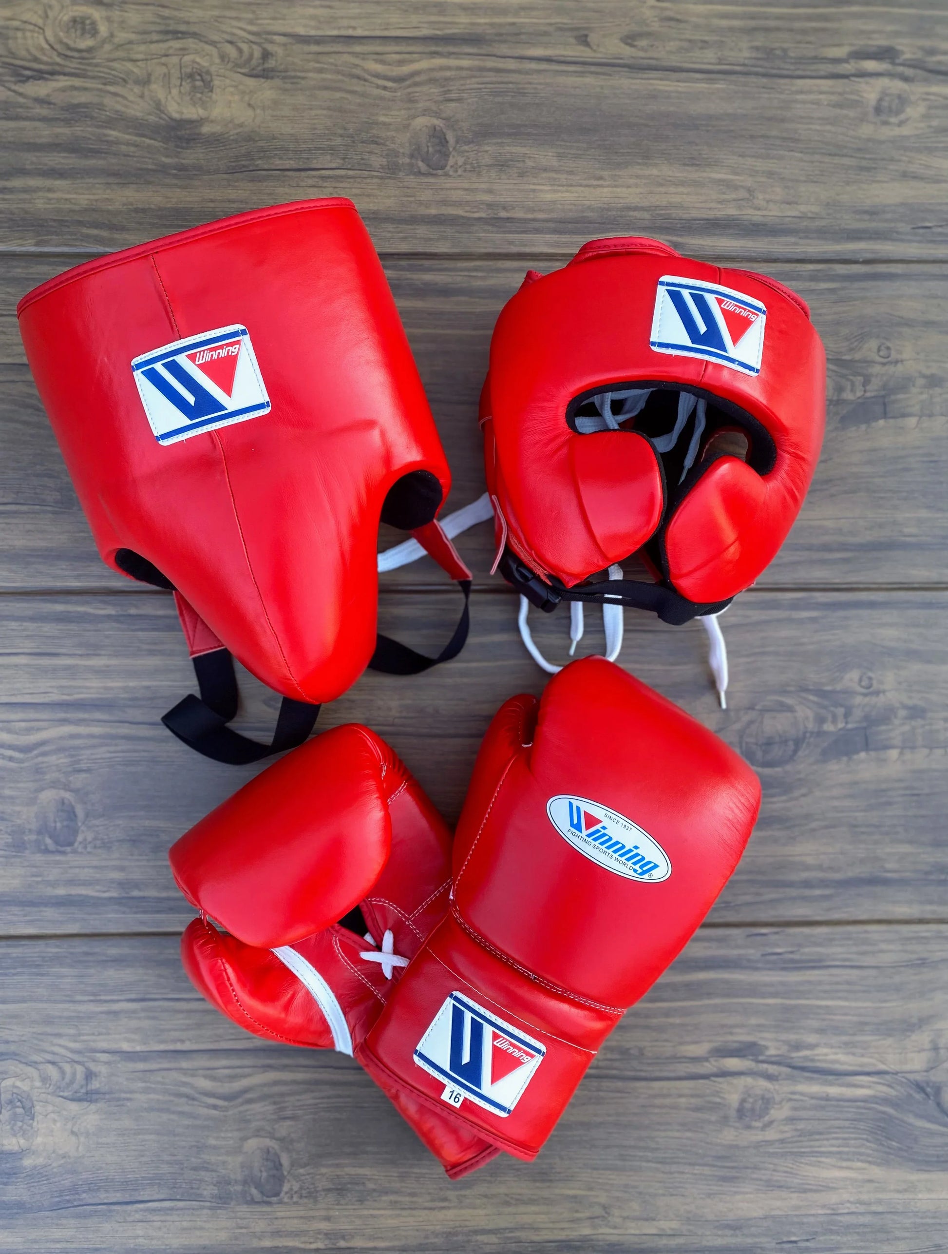 Red boxing gear set including gloves, headgear, and shin guards on a wooden surface.